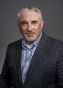 Professional man with short gray hair, wearing a blazer over a checkered shirt, posing confidently against a neutral background named Darragh Heaslip