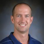 Picture of a smiling man in a blue shirt that has thin white stripes, Travis Bailey