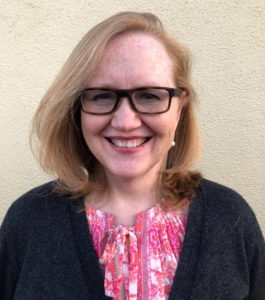 A picture of a smiling woman with shoulder-length blond hair, wearing glasses, a black jacket and a pink floral shirt