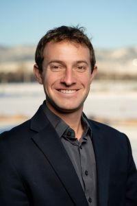 Smiling man in a suit outdoors with mountains in the background, exuding confidence and professionalism.