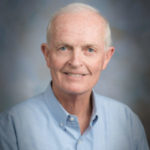 Portrait of a smiling older man in a light blue shirt, set against a soft, blurred background.