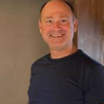 Smiling man in a navy shirt stands indoors, showcasing a friendly demeanor and inviting atmosphere.